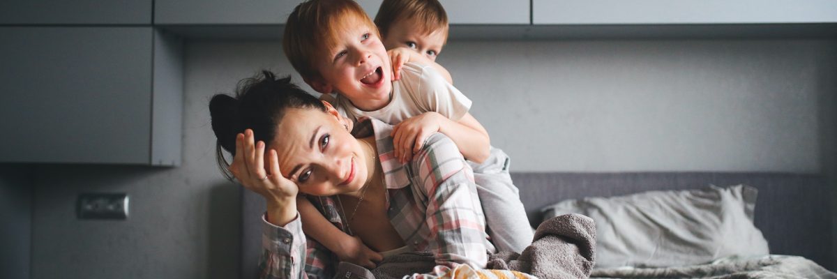 tired mother with two children, crazy children hang on mother in a gray real bedroom. Authentic lifestyle and motherhood difficulties.