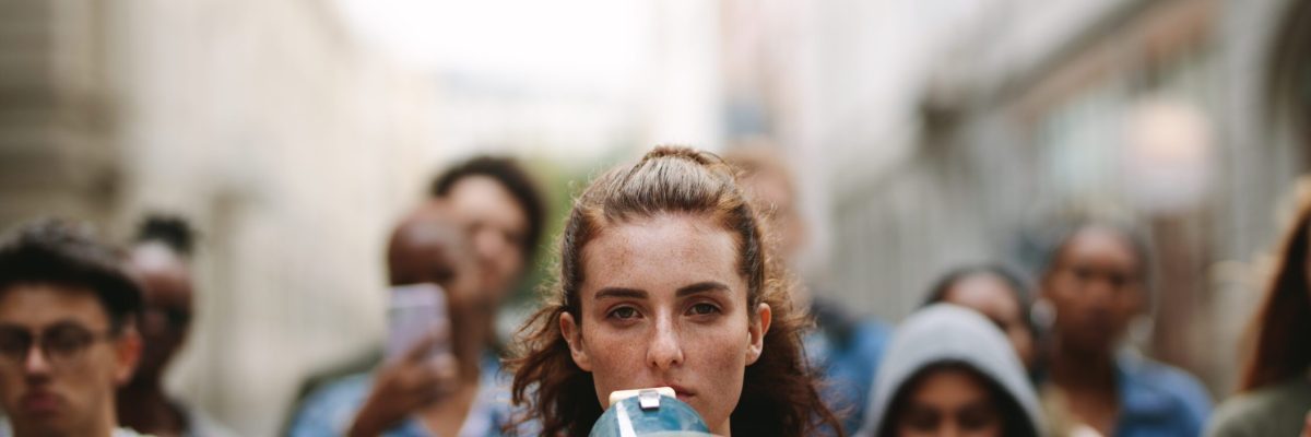 Young woman with a megaphone with group of demonstrator in background. Woman protesting with megaphone in the city.