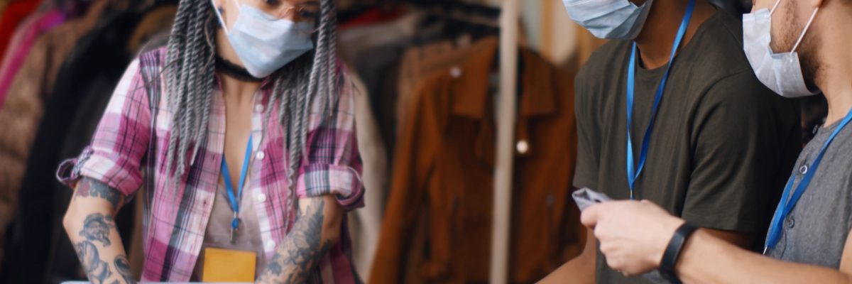 Happy young diverse volunteers in safety mask packing clothes in boxes for charity. Multiethnic people wearing protective mask sorting donation clothes in office