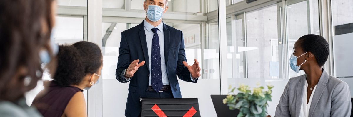 Mature businessman discussing project details with colleagues in modern office with face mask for safety against covid-19 virus. Business people brainstorming and discussing in conference room wearing surgical face mask for covid19 pandemic. Seriuos ceo team talking about the new restrictions due to covid and the social distancing rules.