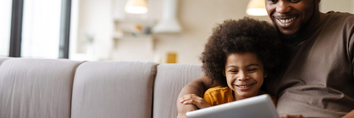 Black father and son smiling and using tablet computer while sitting on sofa at home