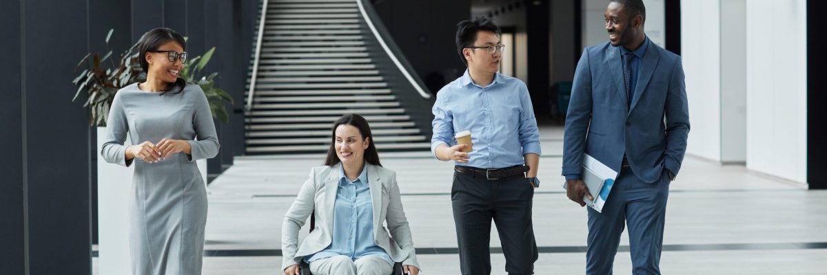 Full length view at diverse group of business people chatting in graphic office lobby while moving towards camera, focus on smiling young woman in wheelchair