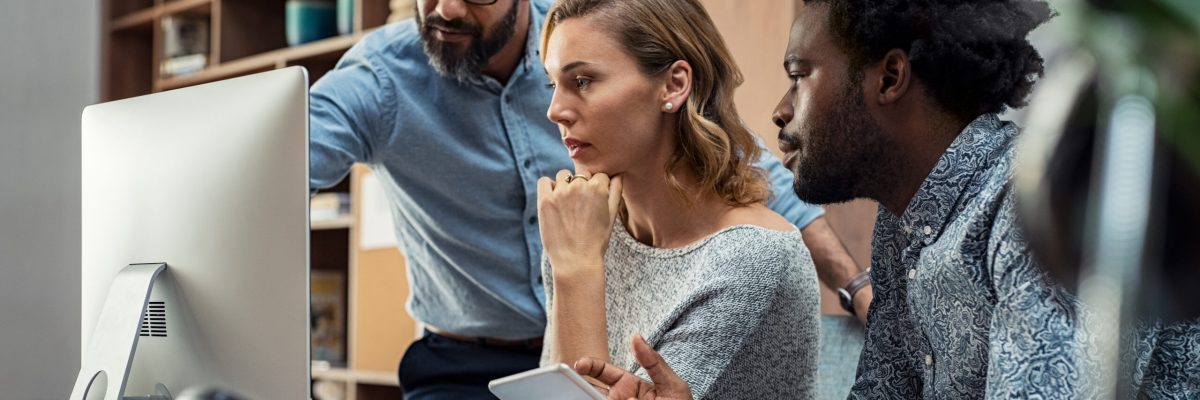 Focused businesspeople discussing project while looking at computer screen. Mature manager working with his creative team in a modern office. Multiethnic casual teamwork at work.